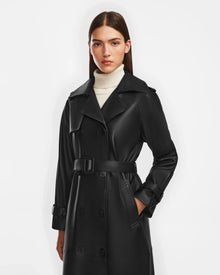 A woman with straight brown hair stands against a white background, wearing the Rudsak CASSIE L - 8124531 black womens trench coat over a white turtleneck sweater.