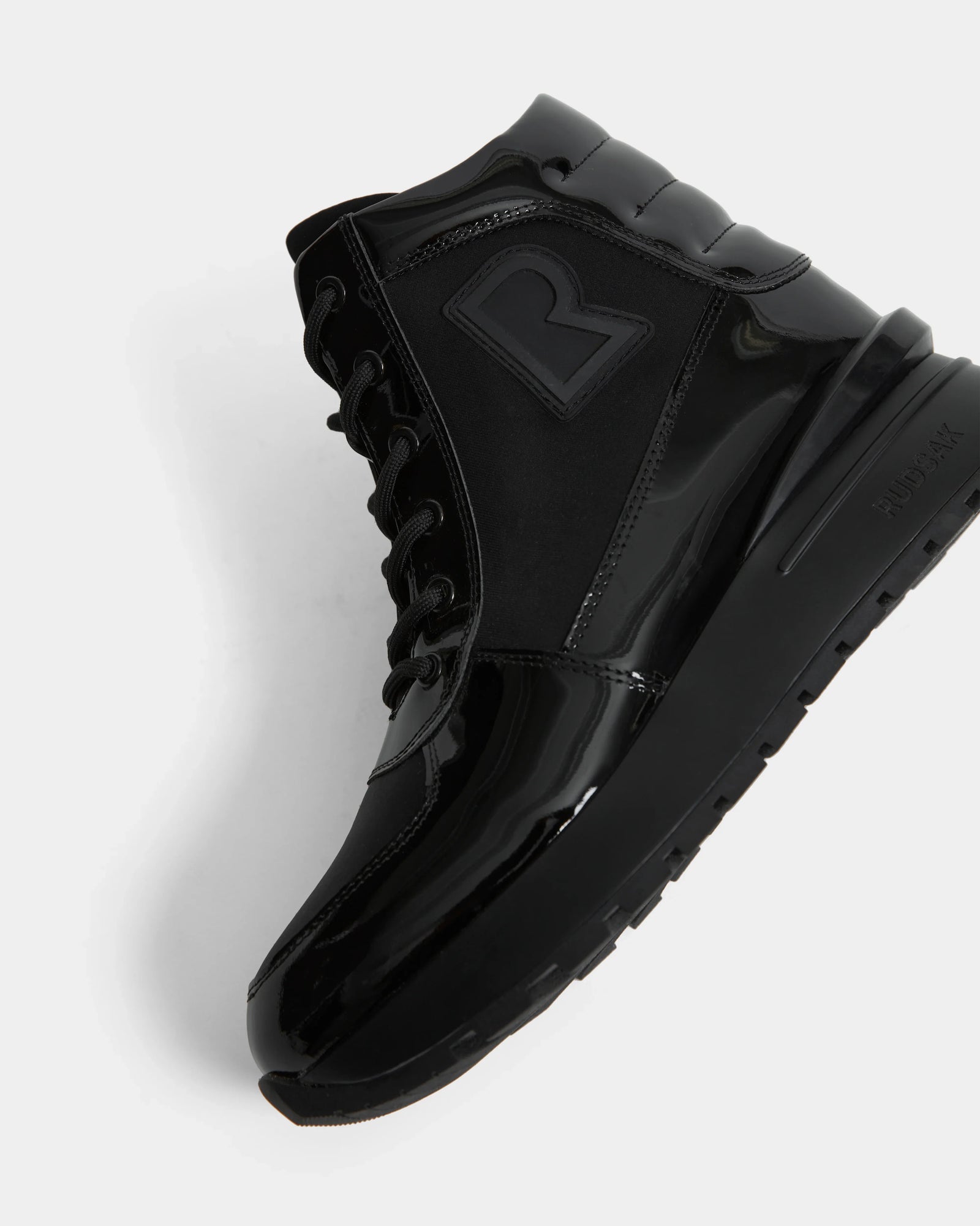The Rudsak CHELSA - L224505 Black is a women’s black high-top sneaker boot with glossy and matte finishes, black laces, a bold “R” side logo, and a waterproof zipper. Shown angled to reveal the side and sole against a white background.