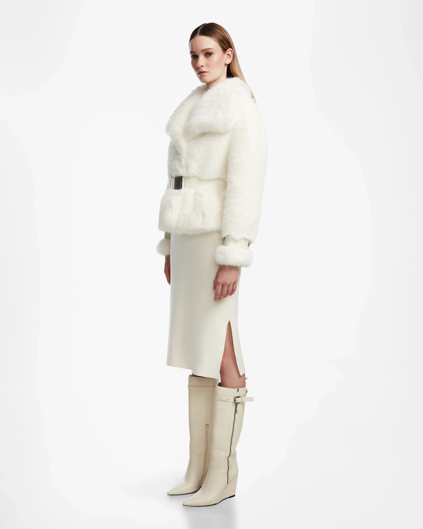 A woman stands sideways wearing the Rudsak CHLOE - L124564 Iceland womens faux fur jacket with belt, a cream midi skirt with side slit, and knee-high vegan leather boots against a plain white background—effortless winter elegance.