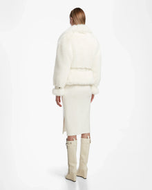 Wearing the Rudsak CHLOE - L124564 Iceland faux fur jacket, a woman with straight, light brown hair pairs it with a white below-the-knee skirt (side slit) and tall cream boots, standing away from the camera—a chic winter wardrobe essential.
