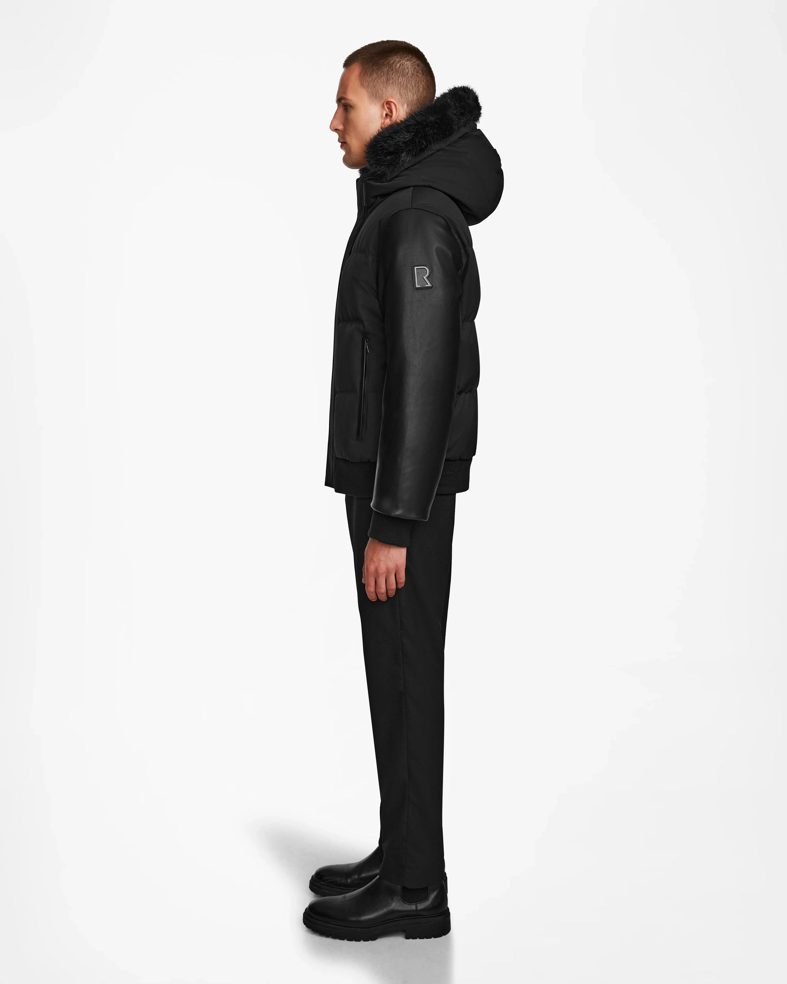 A person stands in profile facing left, wearing the Rudsak DALLAS R - M123501 BLACK weather-resistant jacket with recycled leather sleeves, paired with black pants and black boots against a plain white background.