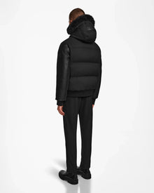 A person faces away, wearing the Rudsak DALLAS R - M123501 BLACK men’s quilted down bomber with recycled leather sleeves and a fur-trimmed hood, paired with black pants and shoes against a plain white background.