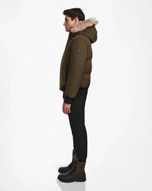 A man stands in profile against a white background, wearing the Rudsak DALLAS - M122501 Army weather-resistant puffer jacket with fur trim, black pants, and brown lace-up boots.