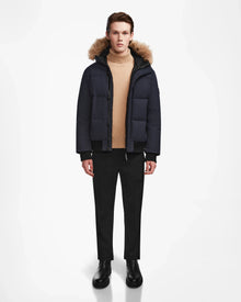 A person faces forward in the Rudsak DALLAS - M122501 Navy men's quilted down bomber with a fur-lined hood, paired with a beige turtleneck, black pants, and black boots against a white background.