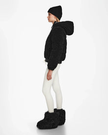 A woman stands in profile against a white background, wearing the Rudsak DANI S - 8124537 Black cropped ski jacket with a fuzzy hood, paired with a black beanie, white leggings, and large black fuzzy boots.