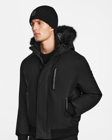 A man wears the Rudsak DEACON - M123513 Black/Black winter coat and black pants, paired with a black beanie, standing against a plain white background and looking slightly to the side.