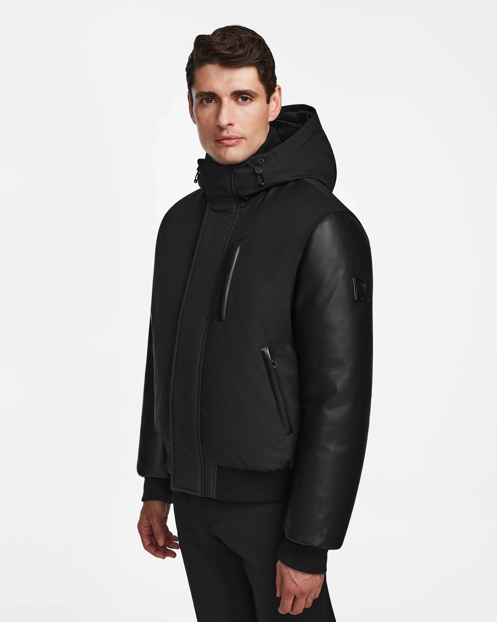 A man with short brown hair wears the Rudsak DERON - M123517 BLACK mens bomber jacket, featuring zippered pockets and weather-resistant material, standing against a plain white background.