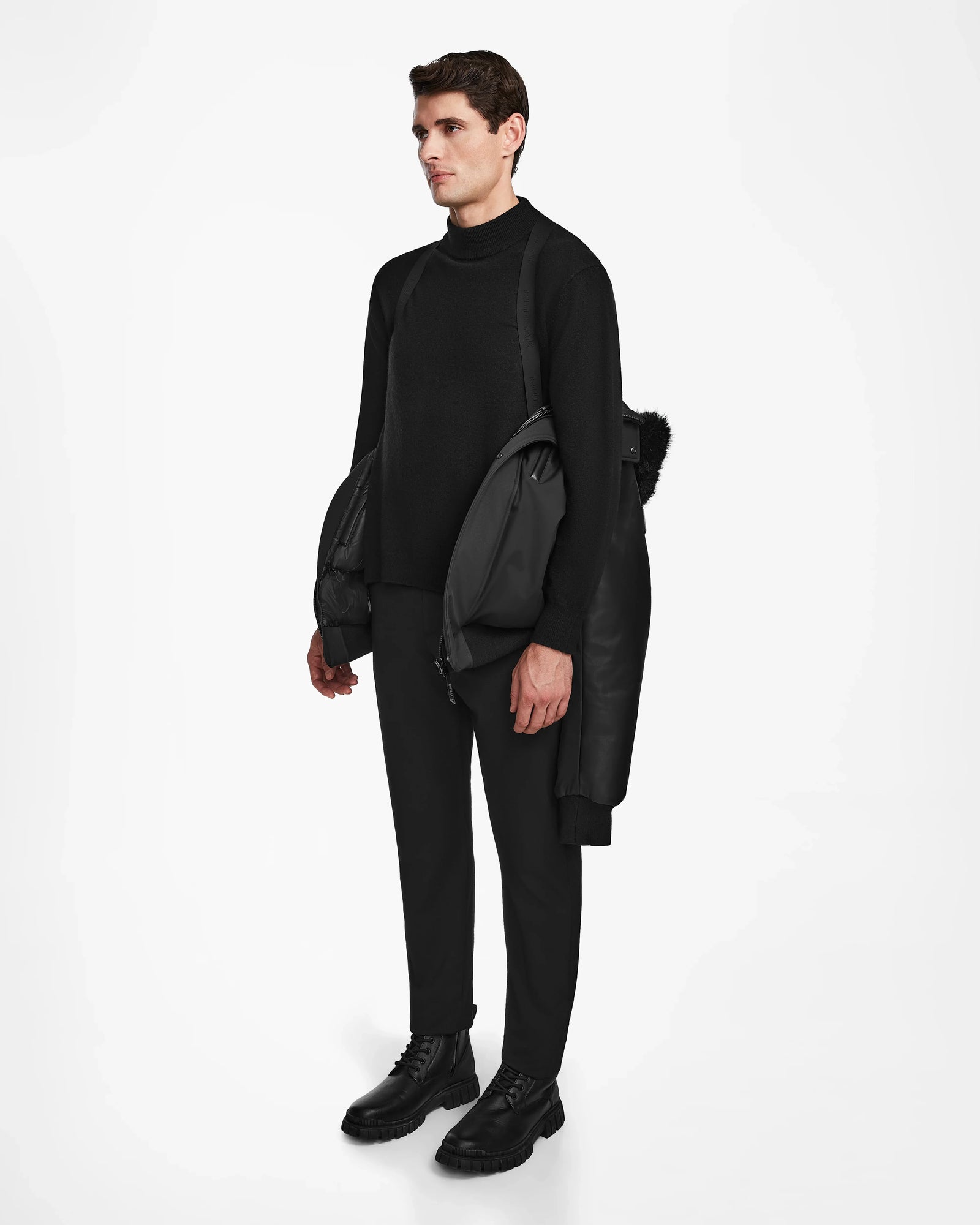 A man stands against a white background in black attire, sporting the weather-resistant Rudsak DERON - M123517 BLACK men’s bomber jacket draped over his shoulders. He gazes to the side, effortlessly blending style and function.