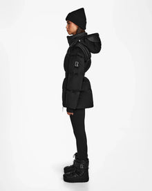 A woman stands in profile against a white background, dressed in the Rudsak DONIA - L124563 Black down puffer coat, black leggings, winter boots, and a black knit beanie.