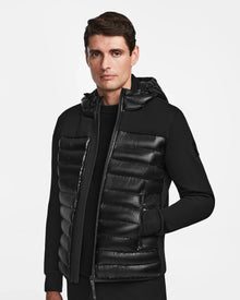 A man wears the Rudsak DURIN S - M123518 Black mens hooded jacket with lightweight down insulation, standing against a plain white background and looking slightly to the side with a neutral expression.