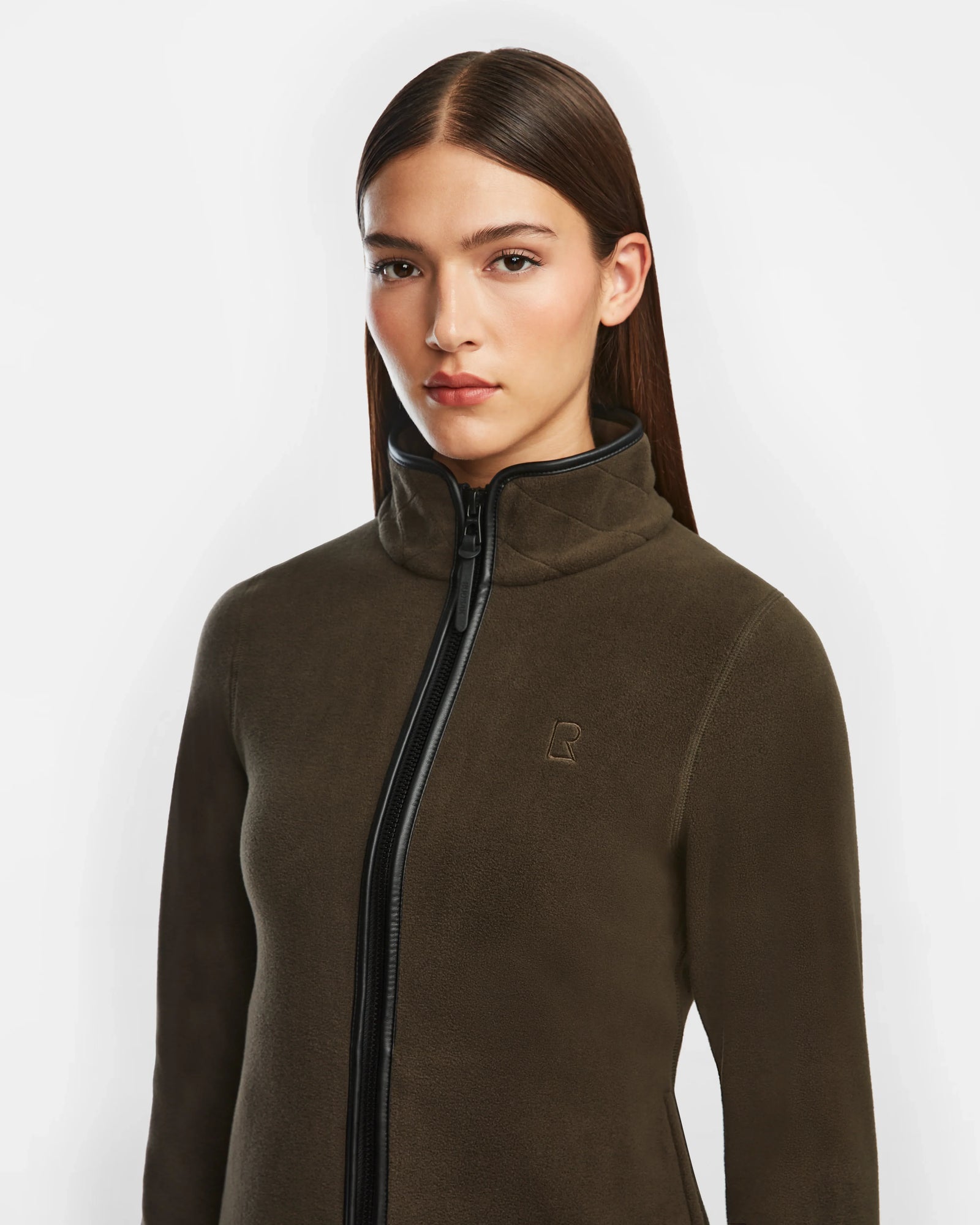 A woman with straight brown hair wears the Rudsak EDITH - 8524617 Army zip-up sweater in dark olive, featuring a high collar, subtle chest logo, soft polar fleece, and vegan leather trim, standing against a plain white background.