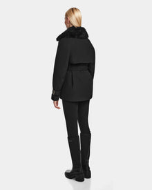 A person with long blonde hair faces away, wearing the Rudsak ELENOR - 8124707 Black womens trench coat with a shearling collar, paired with black leggings and tall black boots against a plain light background.