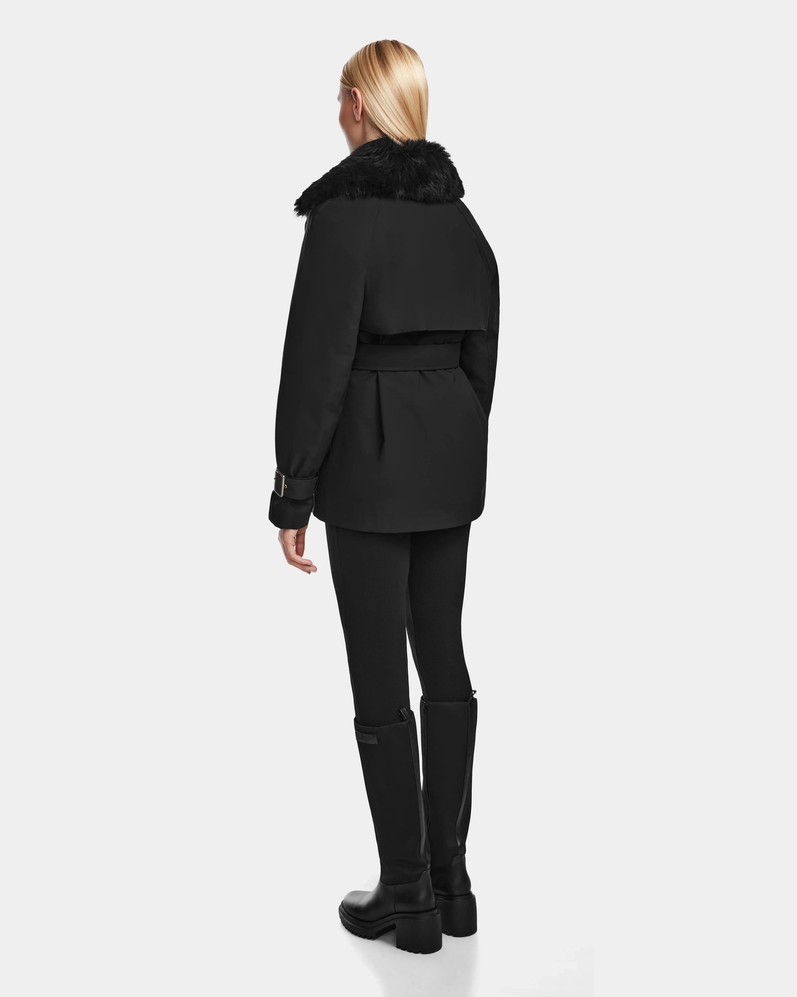 A person with long blonde hair faces away, wearing the Rudsak ELENOR - 8124707 Black womens trench coat with a shearling collar, paired with black leggings and tall black boots against a plain light background.
