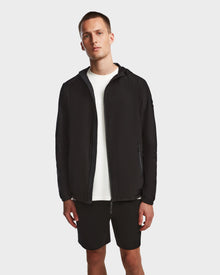 A man stands against a plain background wearing the Rudsak ELIJAH - M124000 Black hooded zip-up jacket over a white t-shirt and black shorts, with his arms relaxed by his sides.