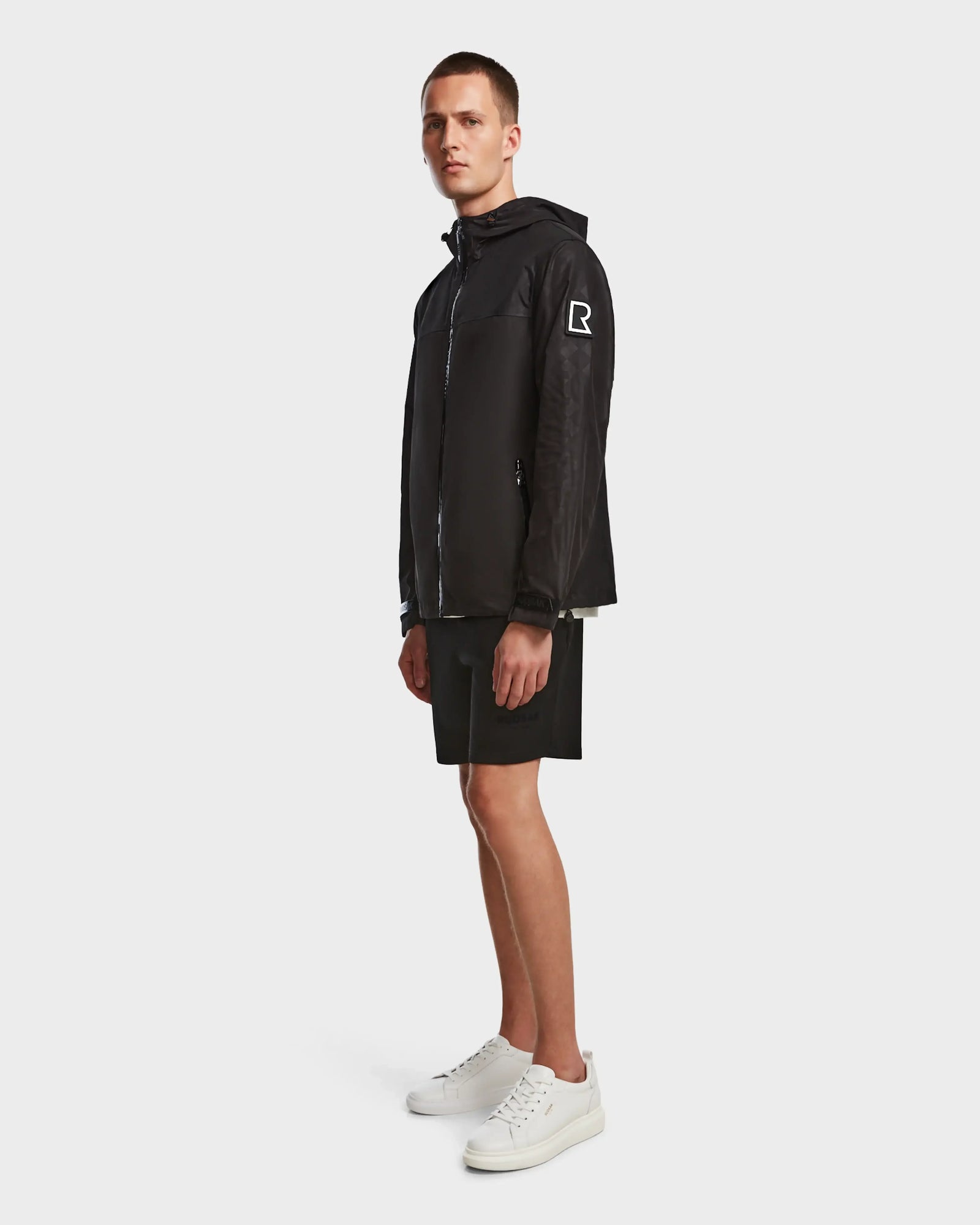 A man stands sideways against a light background wearing the Rudsak ELIJAH - M124000 Black jacket with a white R logo on the sleeve, paired with black shorts and white sneakers.