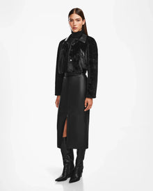 A woman poses against a white background wearing the Rudsak EVELYN - 8124791 Black faux fur jacket, a black leather midi skirt with a front slit, a black top, and pointed black knee-high boots.
