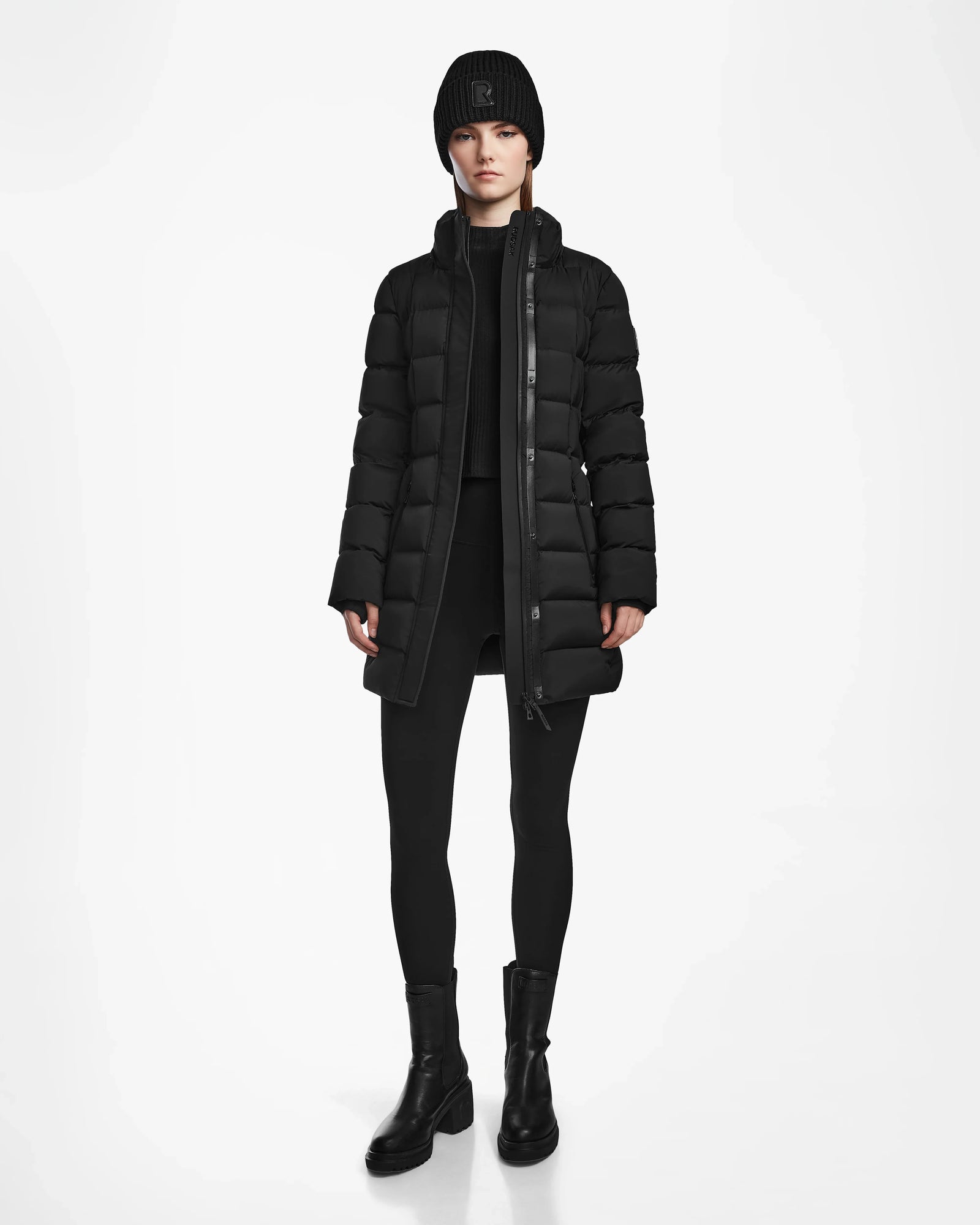 A person stands facing forward against a plain white background wearing the Rudsak FRIDA M - L124524 Black womens mid-length down jacket with a removable hood, paired with a black beanie, black sweater, leggings, and boots.