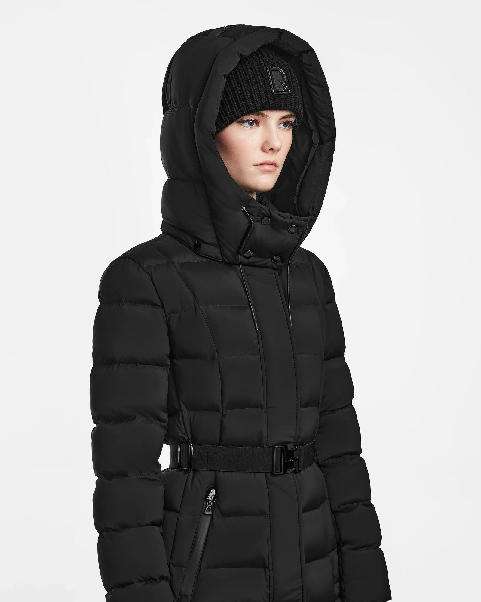 Wearing the Rudsak FRIDA M - L124524 Black quilted down jacket with hood up and a black knit beanie, a person stands against a plain white background, looking to the side with a neutral expression.