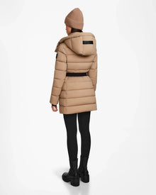 A person is seen from behind wearing the Rudsak FRIDA M - L124524 Sand womens mid-length down jacket with black leggings, black boots, and a beige knit beanie against a white background.