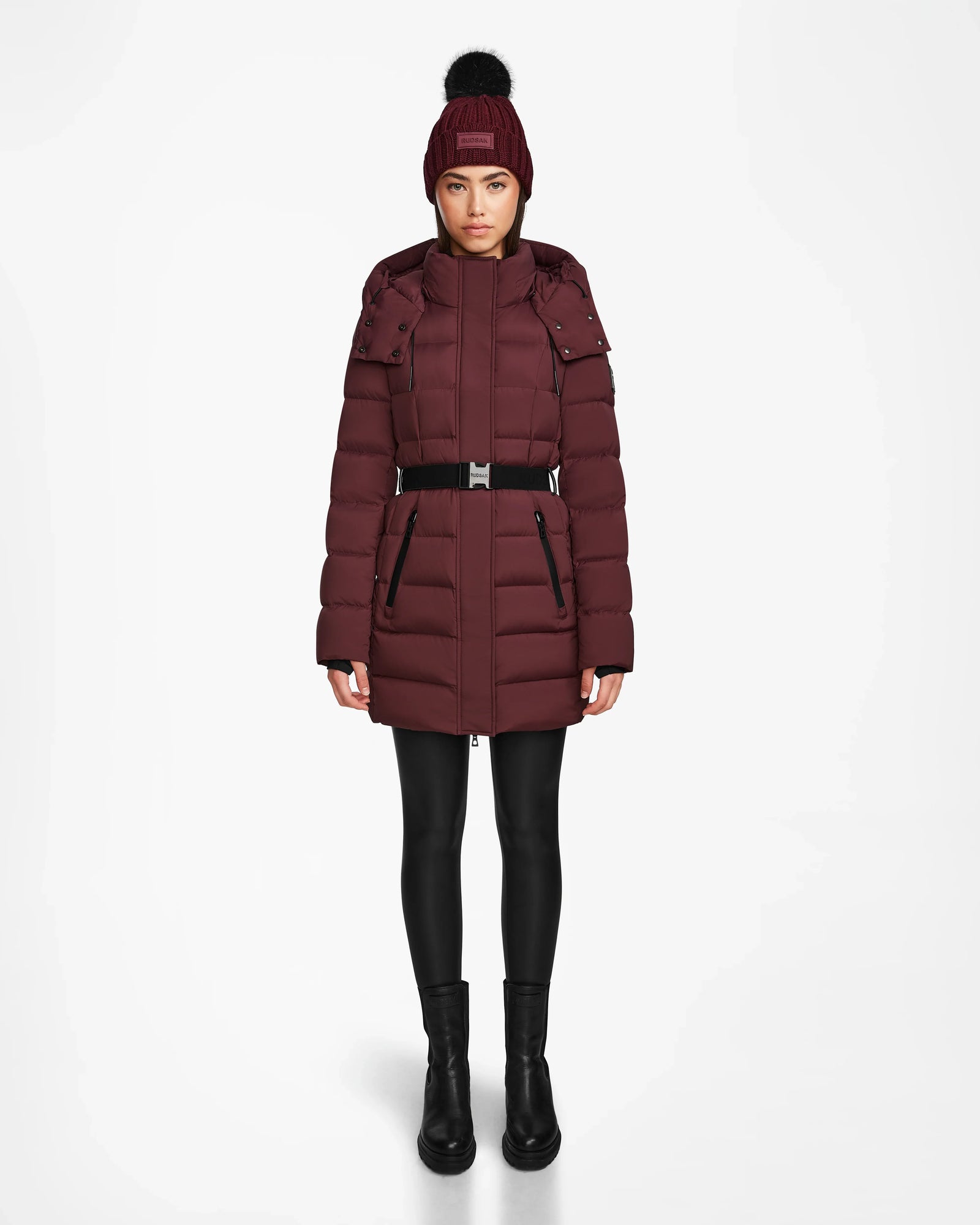A woman faces forward in the Rudsak FRIDA M - L124524 Wine mid-length down jacket with belt, paired with black leggings, boots, and a matching maroon knit hat with black pom-pom, set against a plain white background.