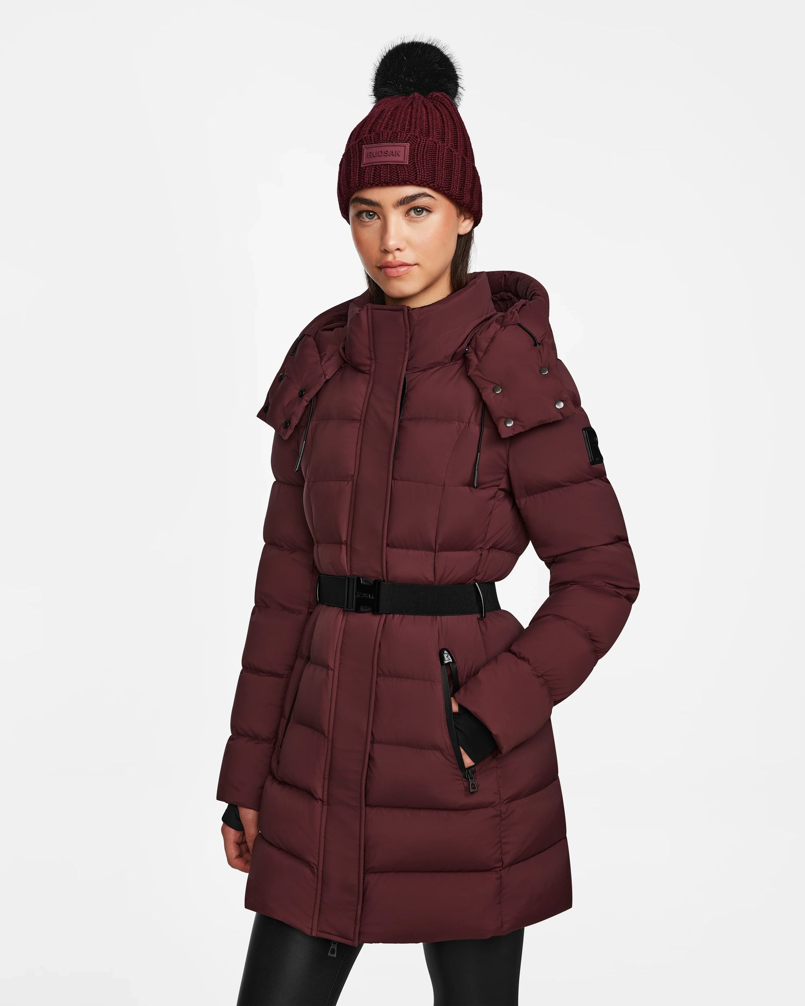 A woman stands against a plain white background wearing the Rudsak FRIDA M - L124524 Wine mid-length down jacket with a belted waist, matching maroon knit beanie, and removable hood.