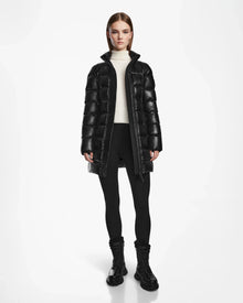 A woman with straight brown hair wears the Rudsak FRIDA S - L124526 Black mid-length down jacket with a removable hood, paired with a white turtleneck, black leggings, and lace-up boots against a plain white background.