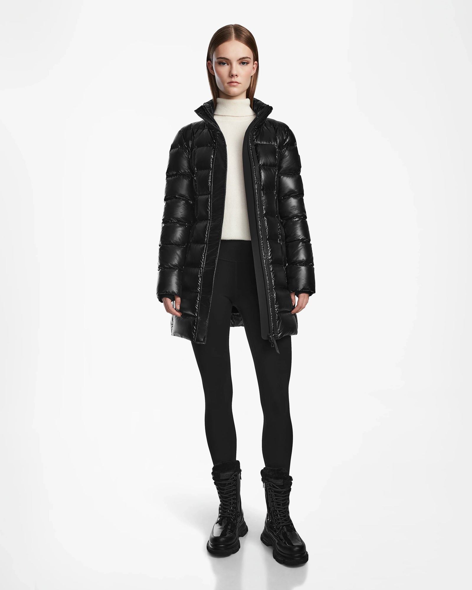 A woman with straight brown hair wears the Rudsak FRIDA S - L124526 Black mid-length down jacket with a removable hood, paired with a white turtleneck, black leggings, and lace-up boots against a plain white background.