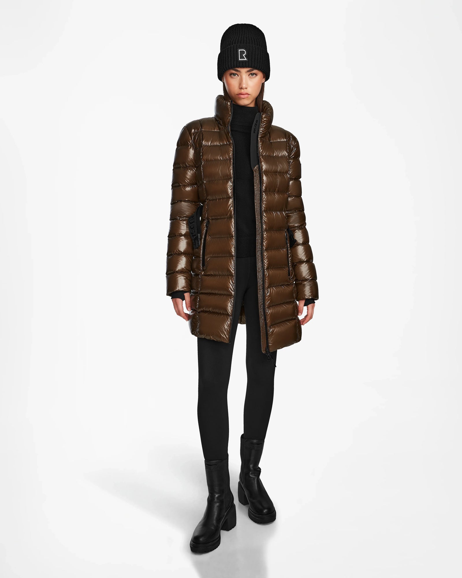 A woman stands facing forward against a plain white background, wearing the Rudsak FRIDA S - L123513 Light Olive shiny quilted down jacket with removable hood, paired with a black knit beanie, turtleneck, leggings, and ankle boots.