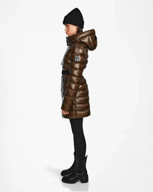 A person stands in profile wearing the Rudsak FRIDA S - L123513 Light Olive women's mid-length down jacket with a hood, black knit beanie, black leggings, and black boots against a plain white background.