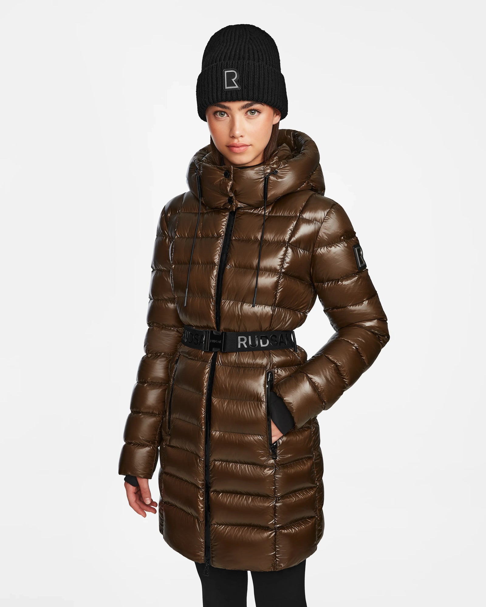 A woman stands against a white background, hands in her pockets, wearing the Rudsak FRIDA S - L123513 Light Olive mid-length down jacket with removable hood, a black beanie with an R logo, and black pants.