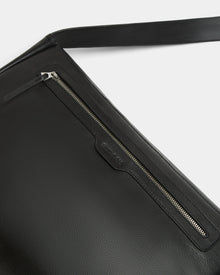 Close-up of the Rudsak GRAYSON L - 8324525 Black leather crossbody bag with a textured finish, silver zipper pocket, and matching leather pull tab. An adjustable strap is partly visible against a white background.
