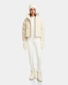 A woman faces forward wearing the Rudsak IKSA - 8124706 Iceland teddy down puffer with a white beanie, matching mittens, fitted white pants, and cream winter boots against a plain light background.