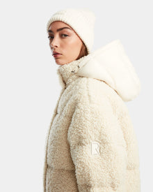 A woman wears the Rudsak IKSA - 8124706 Iceland womens teddy down puffer with hood and matching knit beanie, gazing slightly to the side against a plain light background.