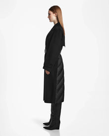 A woman with straight, light brown hair stands in profile wearing the Rudsak IVE - L124560 Black womens wool blend long coat with quilted back detailing and knee-high black boots against a plain white background.