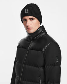A man wearing the Rudsak JACK - M124507 Black down puffer jacket with faux leather accents and a black knit beanie, both displaying the R logo, stands against a plain white background.