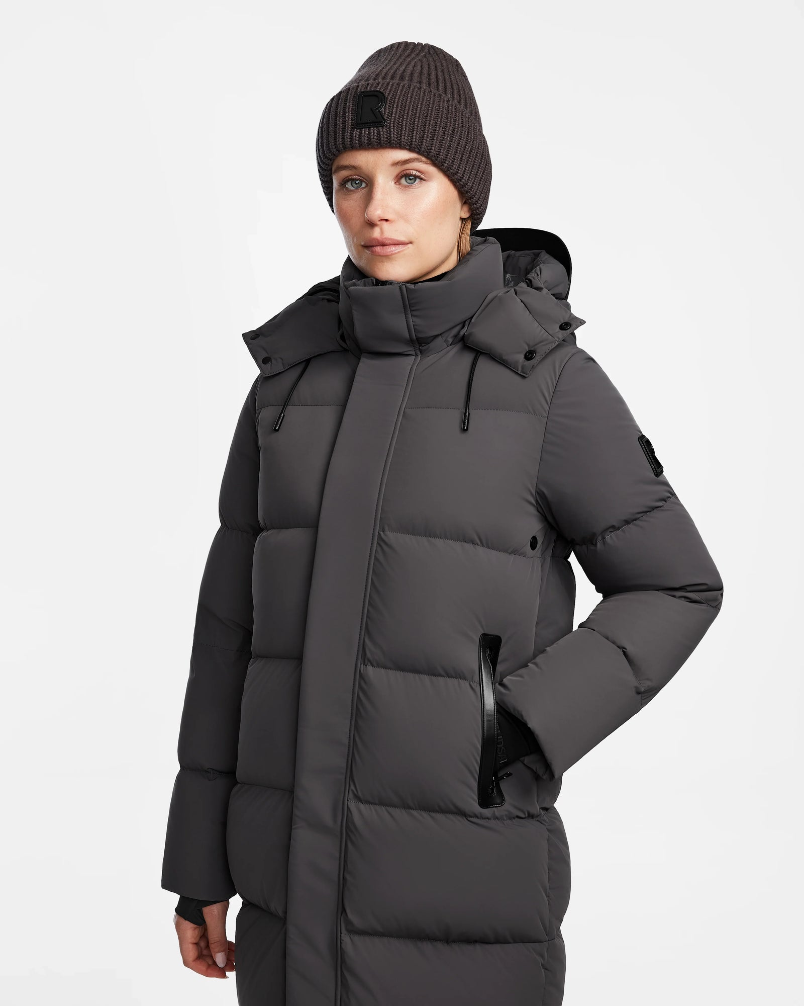 A woman in the Rudsak JOELLE - 8124729 Grey dark gray quilted down coat with a high collar and matching knit beanie stands with one hand in her pocket against a plain white background.