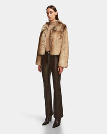 A woman wears the Rudsak JOSILYN - 8124792 Mocha faux fur jacket over a beige top, paired with dark brown leather pants and pointed-toe boots, all set against a plain white background.