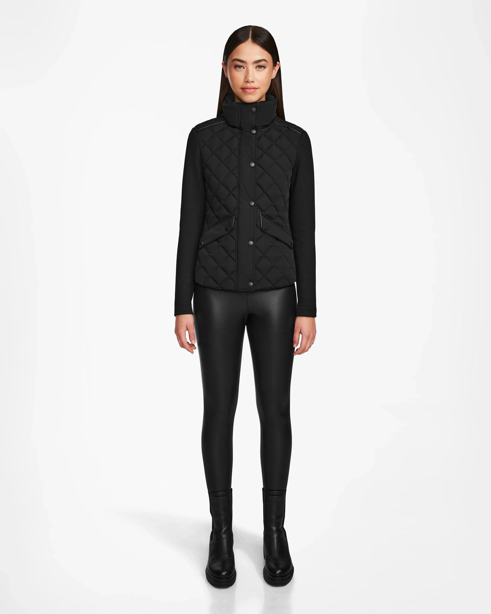 A woman with straight brown hair wears the Rudsak KARMA - L124503 Black matte light jacket with black leggings and boots, standing against a plain white background—ideal outerwear for transitional weather.