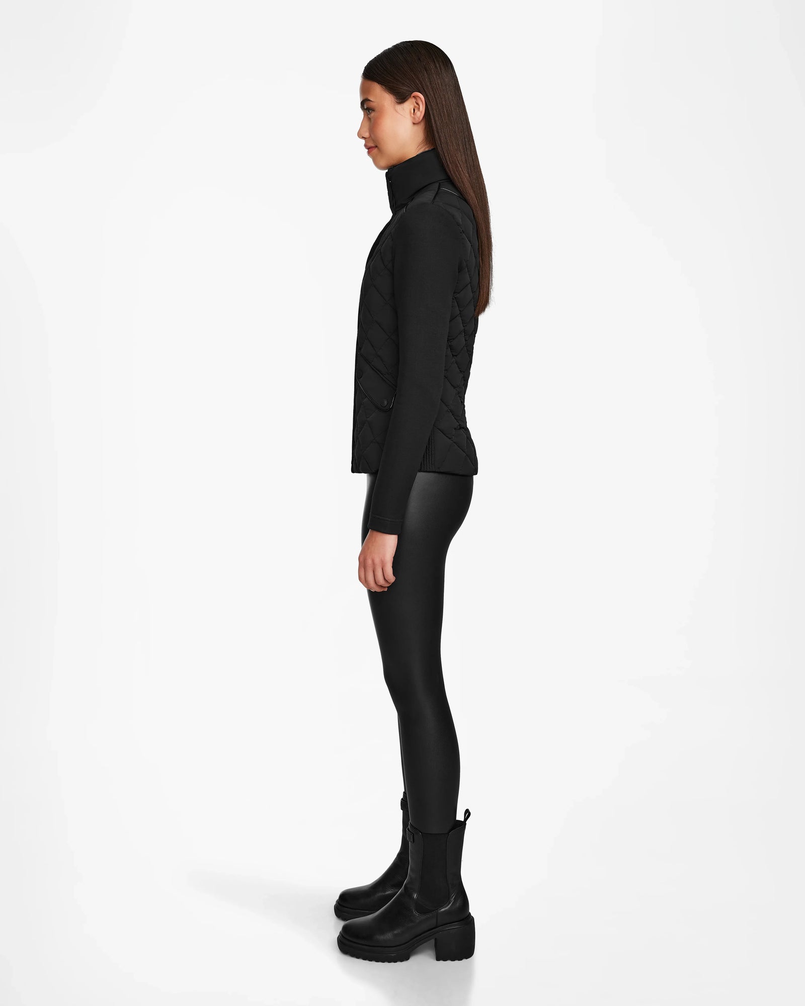 A woman stands in profile against a white background, wearing the Rudsak KARMA - L124503 Black quilted vest, a black long-sleeve top, leggings, and ankle boots—ideal for transitional weather. Her straight brown hair is worn down.