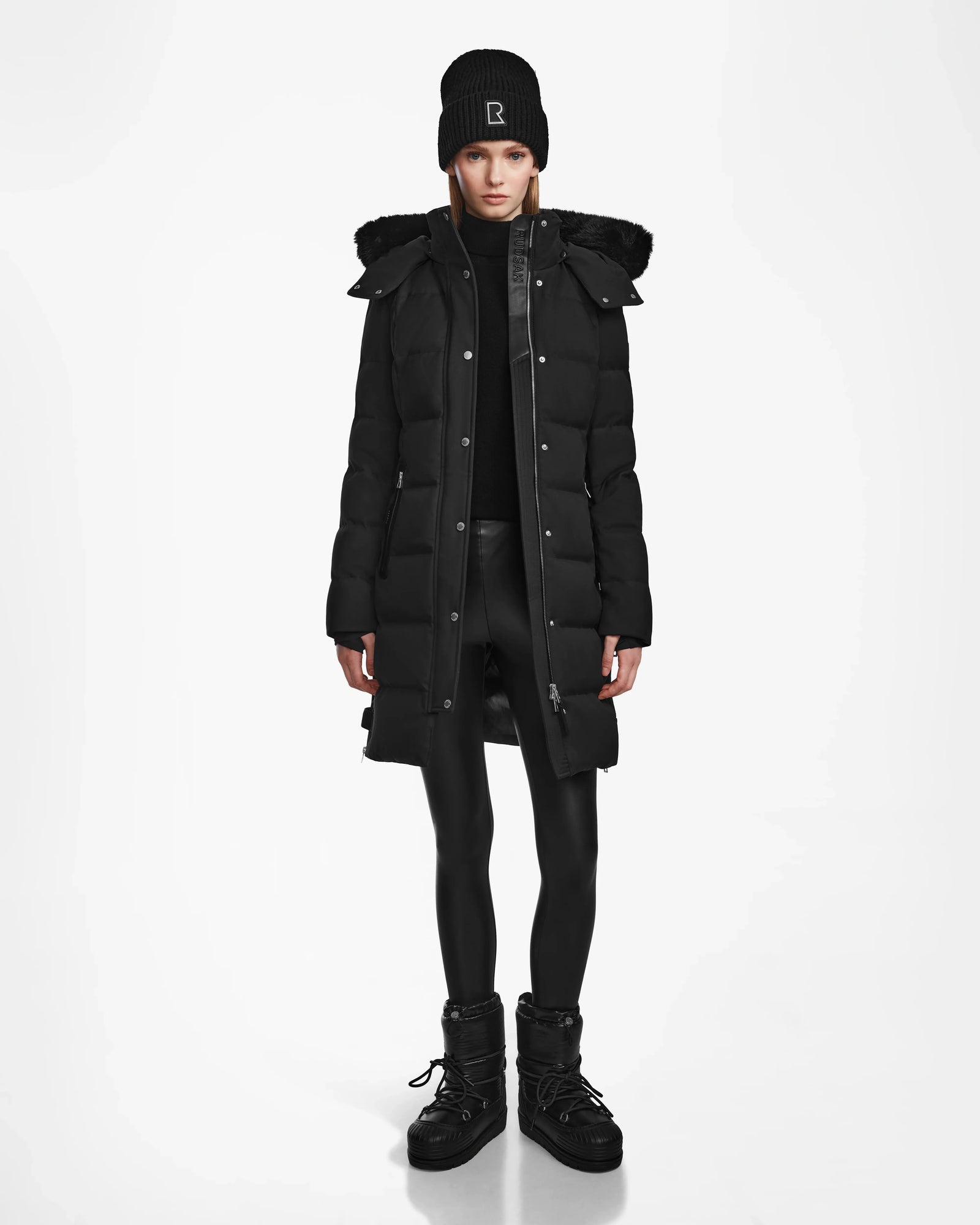 A person stands facing forward against a white background, wearing the Rudsak KEHLANI - L124540 Black down coat with a hood, black beanie, turtleneck, leggings, and boots—a true winter essential.