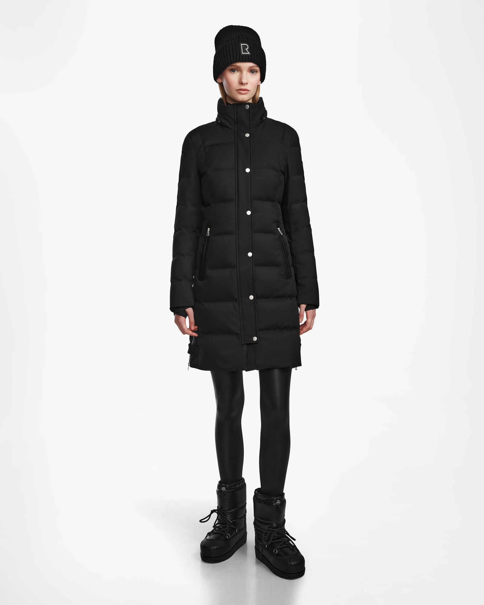 A person faces forward against a plain white background, wearing the Rudsak KEHLANI - L124540 Black long quilted down coat, paired with a black knit beanie, black leggings, and black winter boots.