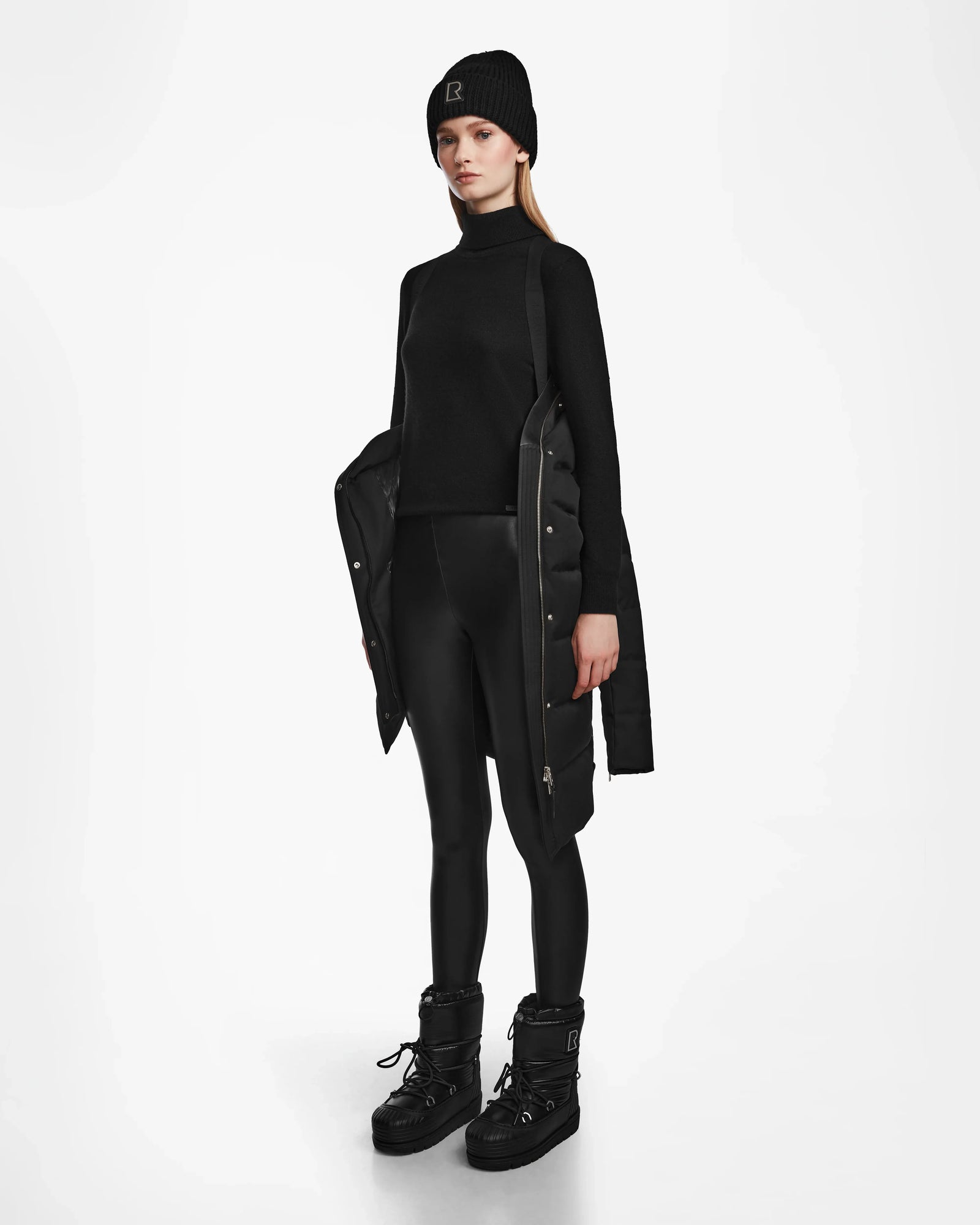 Dressed in the KEHLANI - L124540 Black womens down coat by Rudsak, paired with a black beanie, turtleneck, shiny leggings, and bulky boots, this look is the epitome of winter essential style against a white background.