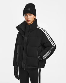 A woman wearing the Rudsak KIMIA - 8124716 Black ski jacket with white sleeve stripes, black leggings with a white stripe, a black beanie, and black gloves stands against a plain white background.