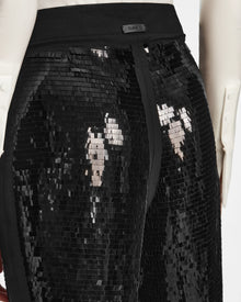 A close-up of a person wearing Rudsak’s LEAR - 8524553 Black high-waisted sequin pants with a black waistband, paired with a white long-sleeve shirt featuring structured cuffs. The shiny sequins give these trousers a festive, eye-catching look.