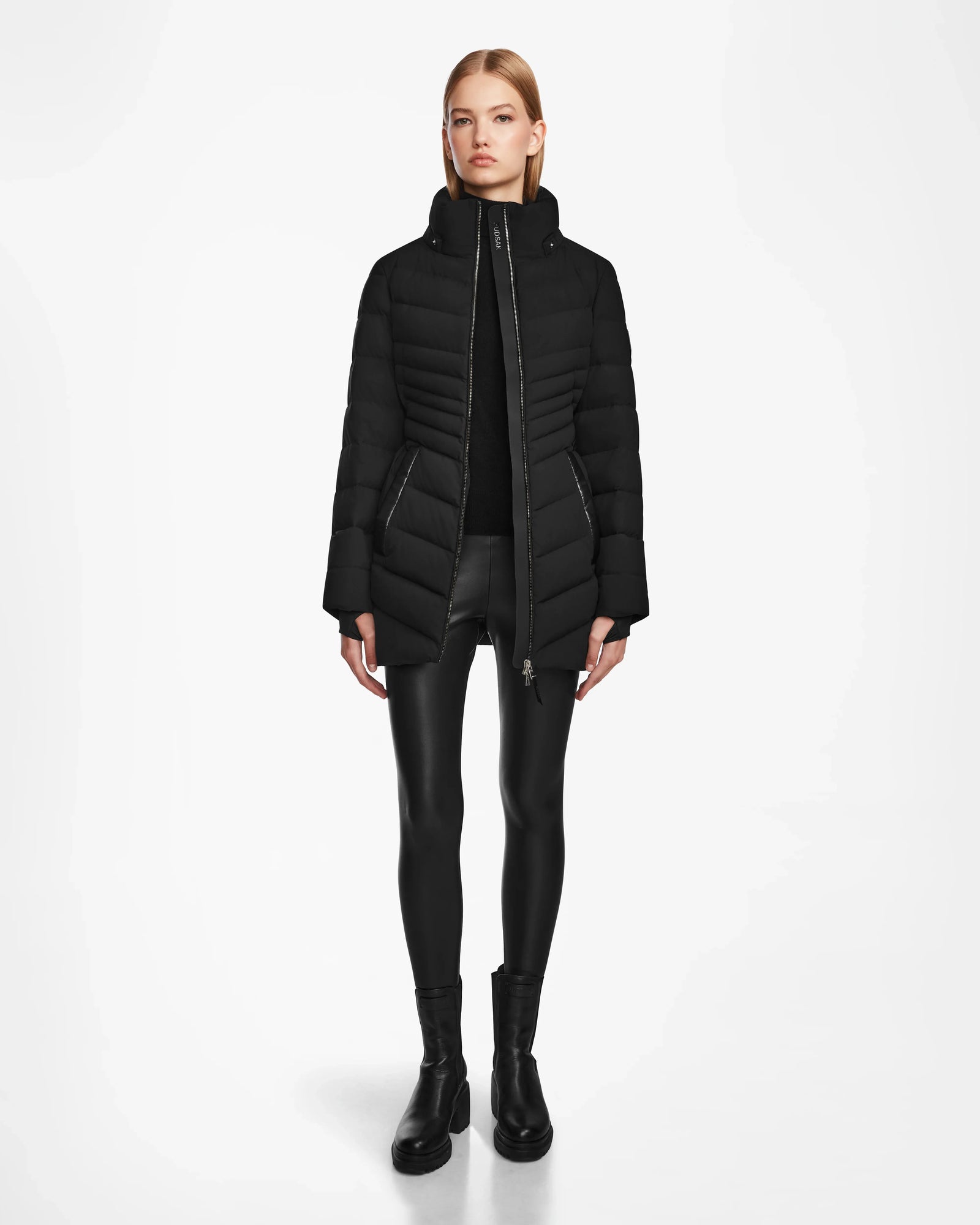 A woman faces forward against a white background, wearing the Rudsak MALAYA M - L123554 Black womens down puffer jacket with a sleek herringbone quilting, paired with a black top, leather-look leggings, and ankle boots.