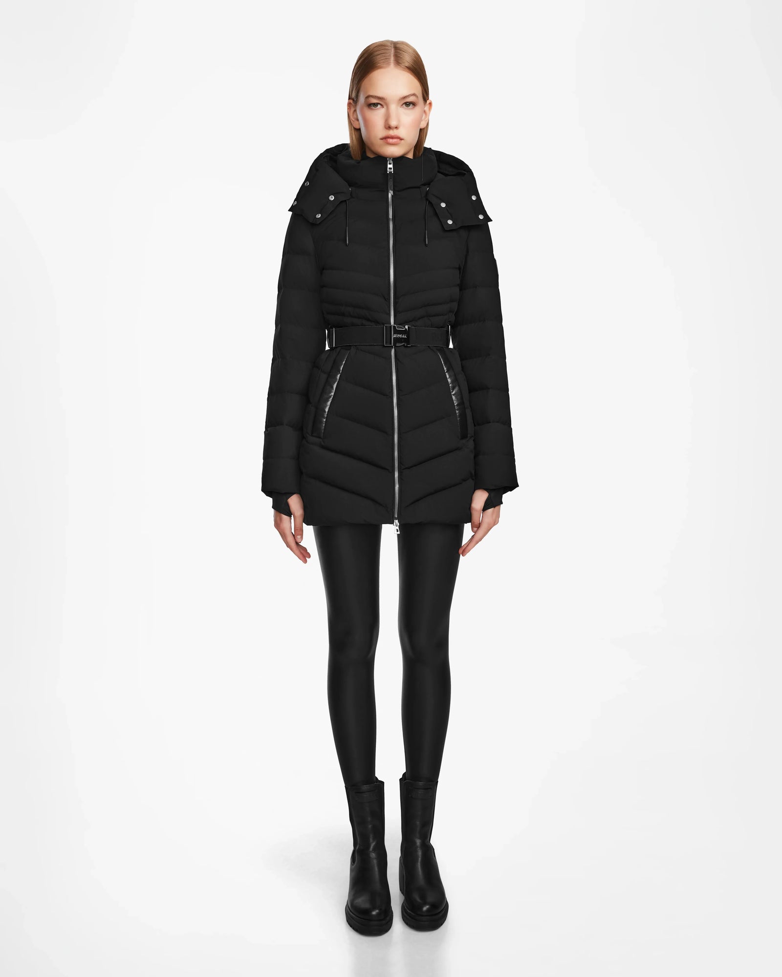 A woman faces forward against a white background wearing the Rudsak MALAYA M - L123554 Black women’s down puffer jacket with a hood, belted waist, and herringbone quilting, paired with black leggings and ankle boots.