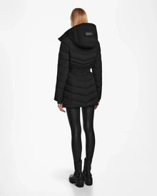 A woman with straight, blonde hair stands facing away, wearing Rudsak’s MALAYA M - L123554 Black women’s down puffer jacket with herringbone quilting, shiny black leggings, and ankle boots against a plain white background.