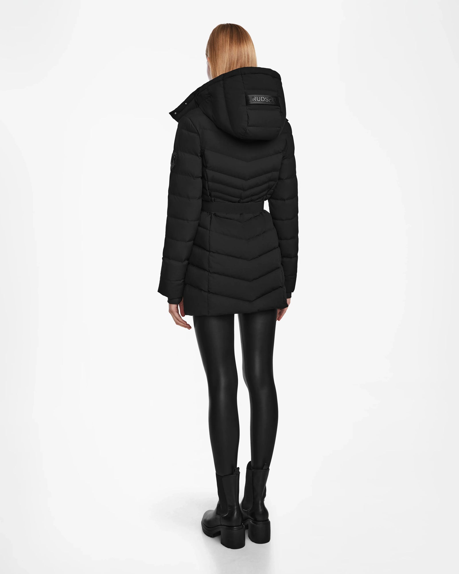 A woman with straight, blonde hair stands facing away, wearing Rudsak’s MALAYA M - L123554 Black women’s down puffer jacket with herringbone quilting, shiny black leggings, and ankle boots against a plain white background.