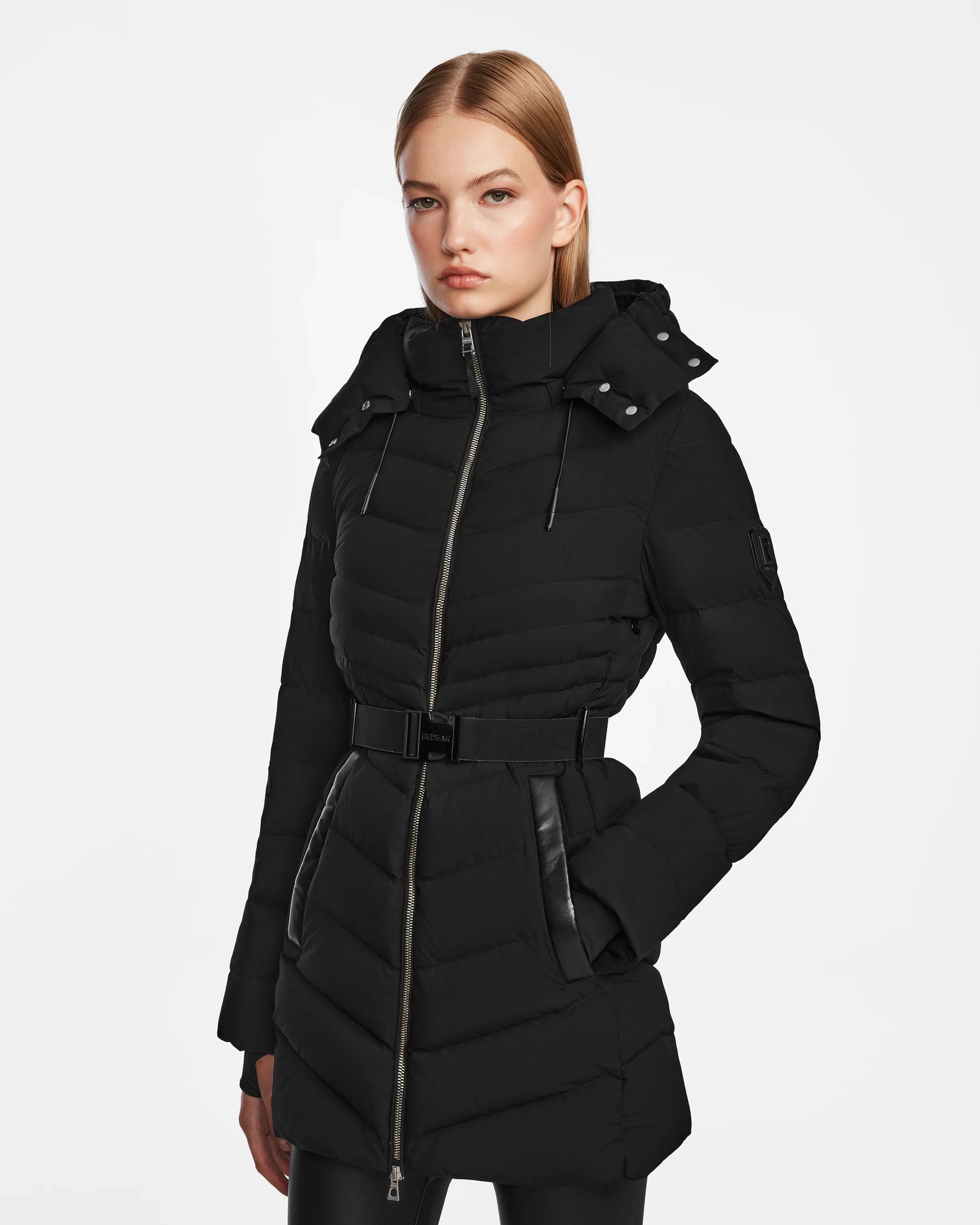 A woman with straight blonde hair models the Rudsak MALAYA M - L123554 Black women’s down puffer jacket, featuring a hood, zip front, belted waist, leather accents, and subtle herringbone quilting against a white background.