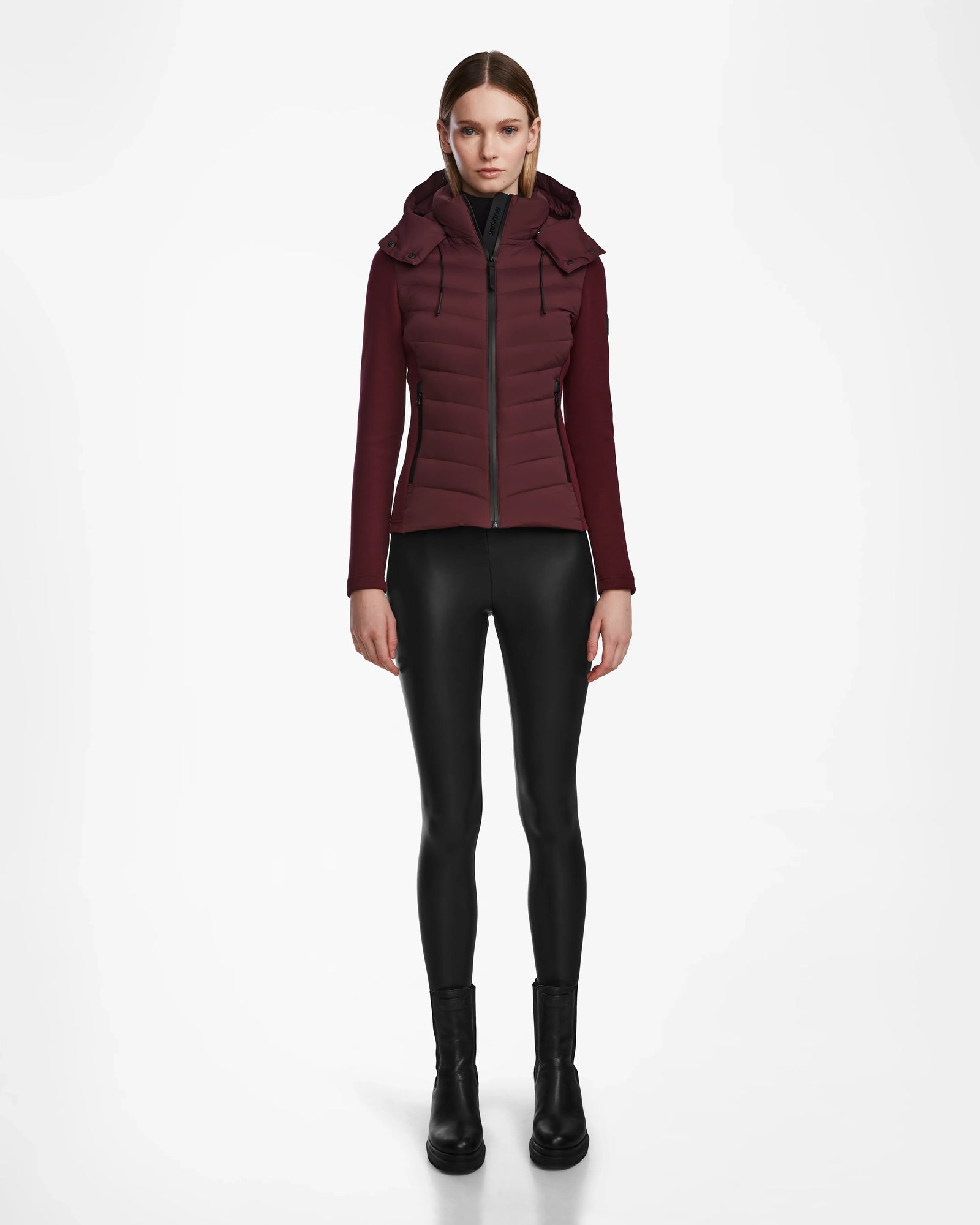 A woman stands facing forward, wearing the Rudsak MALYA M - L123547 Wine down quilted jacket, black shiny leggings, and black boots against a plain white background.